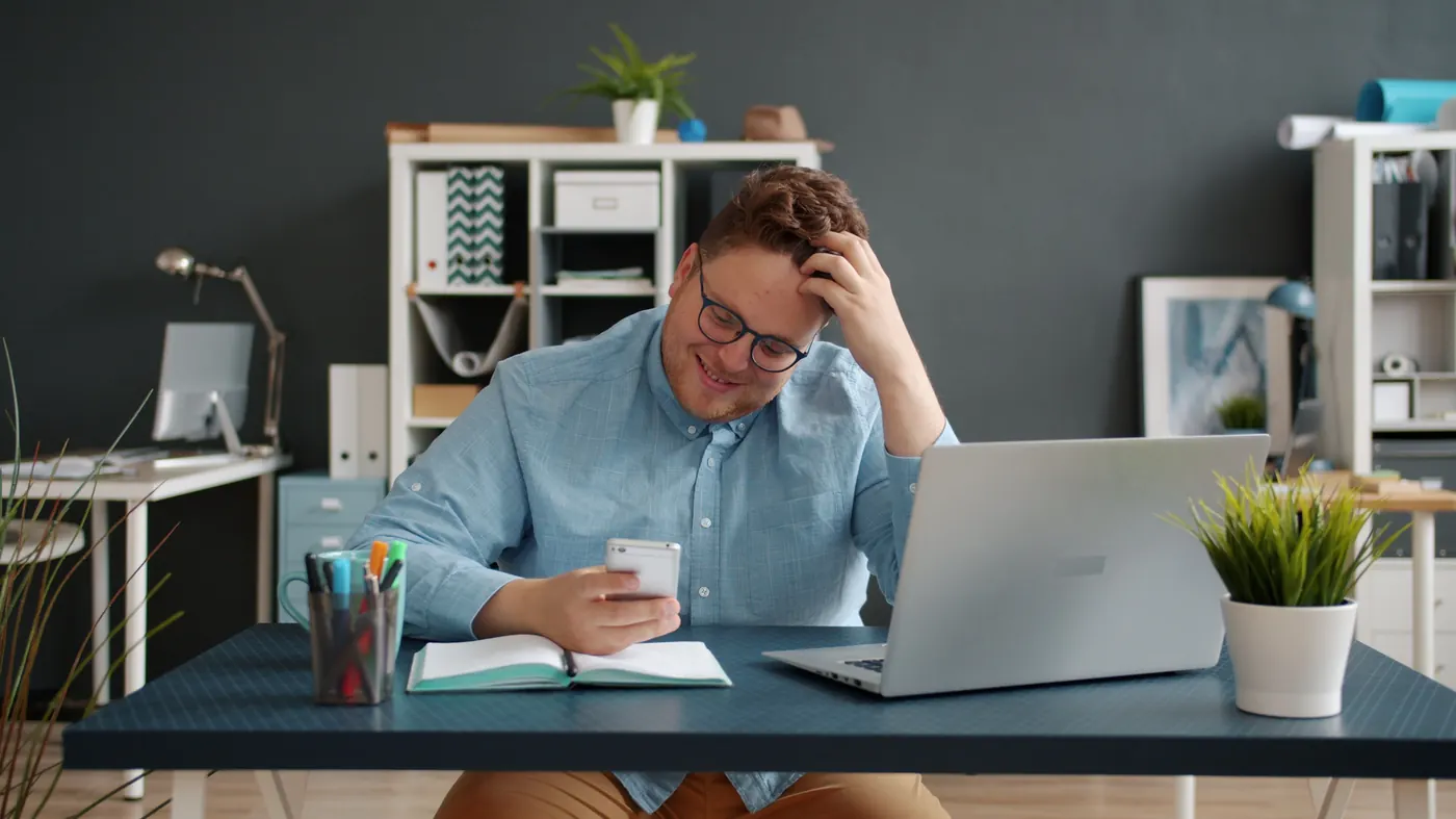 Homem sentado à mesa de trabalho, com notebook aberto, olhando para o celular em vez de trabalhar